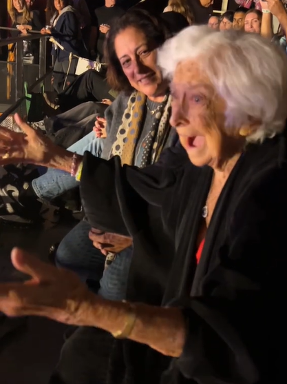 Rachel Platten's 100-year-old neighbor, Gloria, sits in the audience at one of her shows. Gloria's mouth is agape and her eyes are wide as she holds her arms out in front of her, shocked.