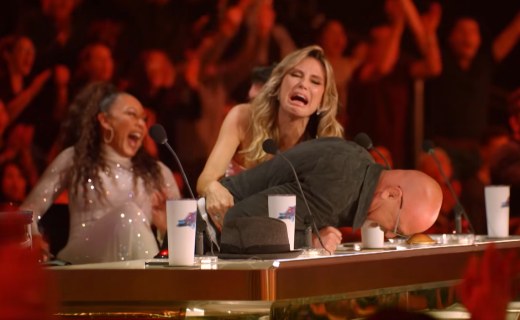 Heidi Klum makes an exagerated crying face as she tries to pull back Howie Mandel who is hitting the Golden Buzzer with his forehead. Mel B. laughs in the background. 