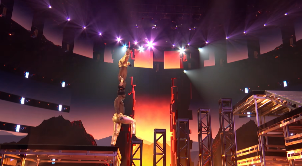 Side-view of the Ramadhani Brothers performing on "AGT." One of them is blindfolded as he takes the first step onto a rising staircase with six steps. The second is balancing on top of his head on his head.