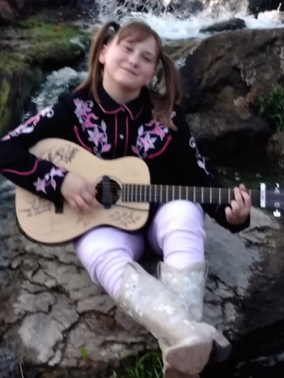 Ruby Leigh, when she was a child, smiles as she sits on a large rock. A waterfall is in the distance. She's wearing a cowgirl-style shirt and boots. She smiles as she plays a guitar.