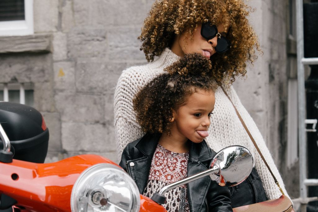 A woman and her little girl pose next to a motorcycle. They're both sticking their tongues out as they look at themselves in the motorcycle's mirror.