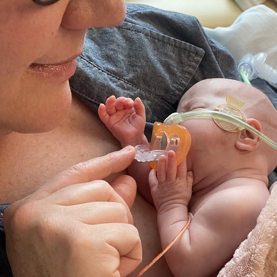 Close up of Ashley McClinton smiling as she hold her baby, John, in her arms. John has grown. He has a pacifier in his mouth and is on oxygen.