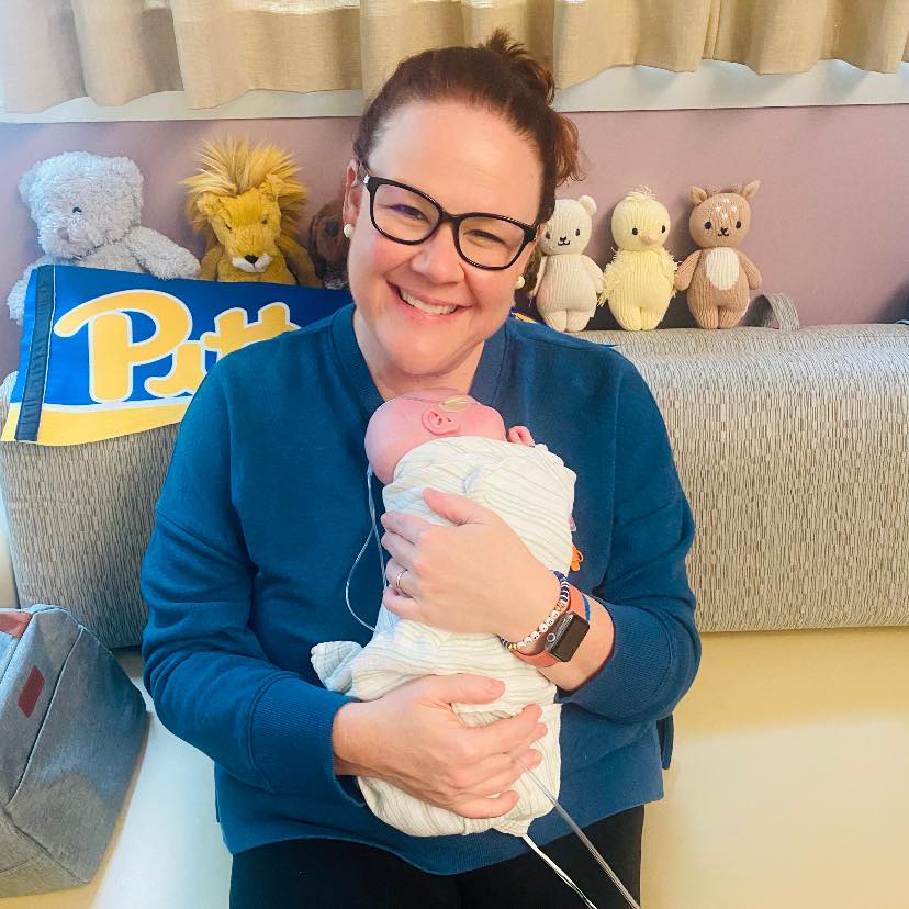 Ashley McClinton smiles wide as she sits on a couch and holds her swaddled baby, Joh, in her arms.