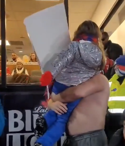 Behind shot of Jason Kelce lifting a young girl holding a sign. He's lifting her to get a better view of Taylor Swift who is in a box at the Chiefs-Bills game.