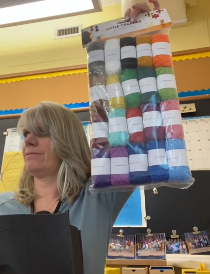 A teacher holds up a large, clear bag full of 20 skeins of color yarn, showing them off to her class.