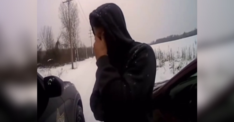 A man wearing a hoodie stands outside on the side of the road. Snow is on the ground. One one side of him is his own car and on the other is a cop car. The man covers his face with his hand as he cries.