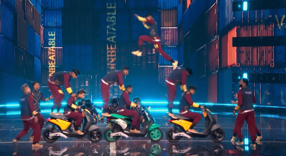 Three men on motorcycles drive across the "AGT" stage, each with a person standing on the back and leaning on the driver for support. Above them, mid-air, a man leaps off the back of one person on a motorcycle to get to the next one. These are members of the dance group V.Unbeatable. 