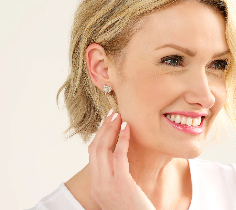 A woman looks off in the distance as she smiles and puts a hand close to her ear. She's wearing Affinity Diamonds Heart Stud Earrings.