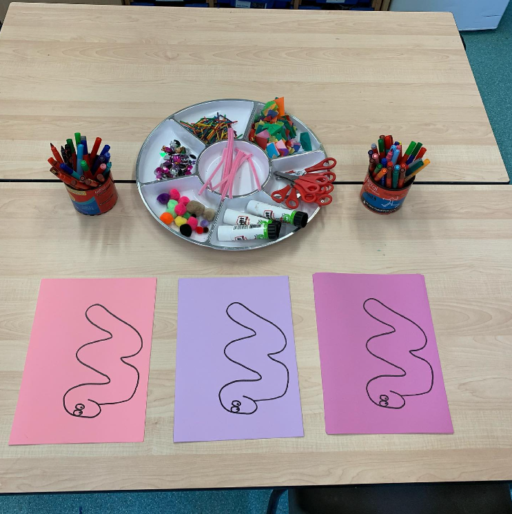 Arts and crafts table with drawings of three worms on three papers: pink, blue, and purple. There are scissors, pop-poms, googly eyes, and more to decorate them.
