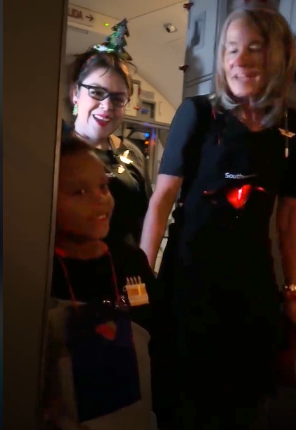 A little boy smiles as two flight attendants stand with him at the front of a plane, singing "happy birthday" to him.