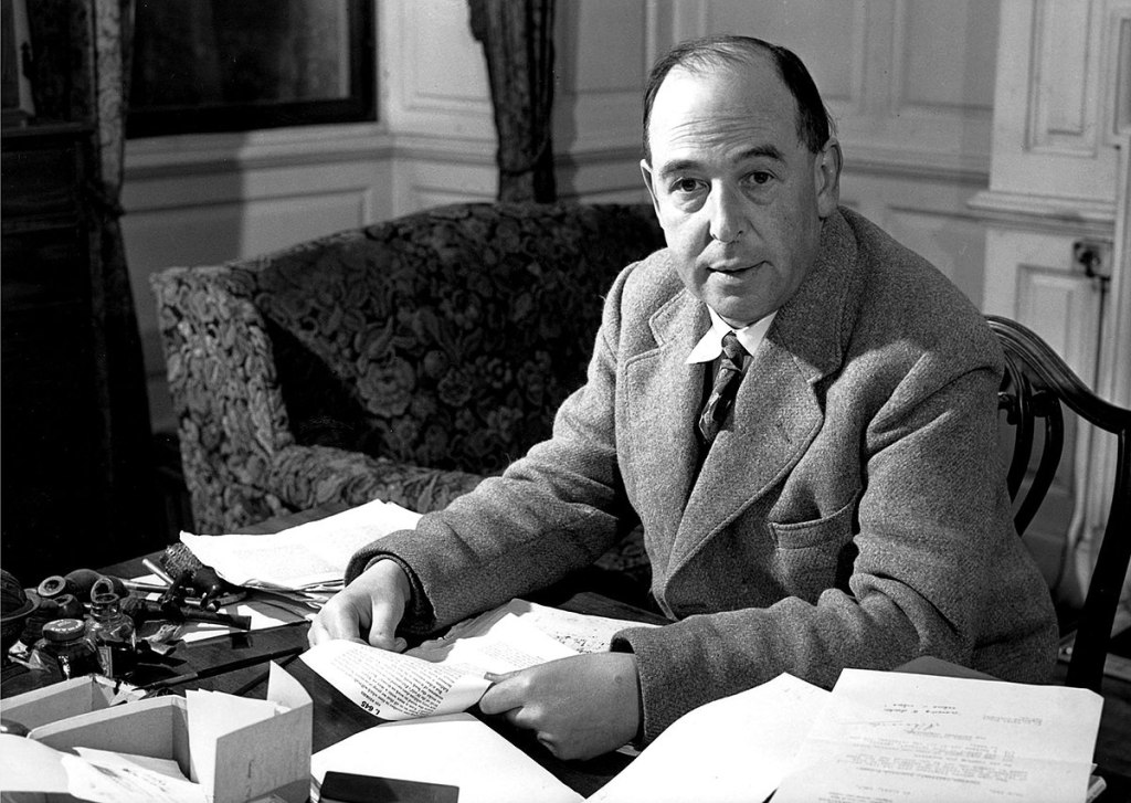 Black and white photo of C.S. Lewis sitting at his desk. He's holding onto a piece of paper, but there's scattered papers all over the desk.