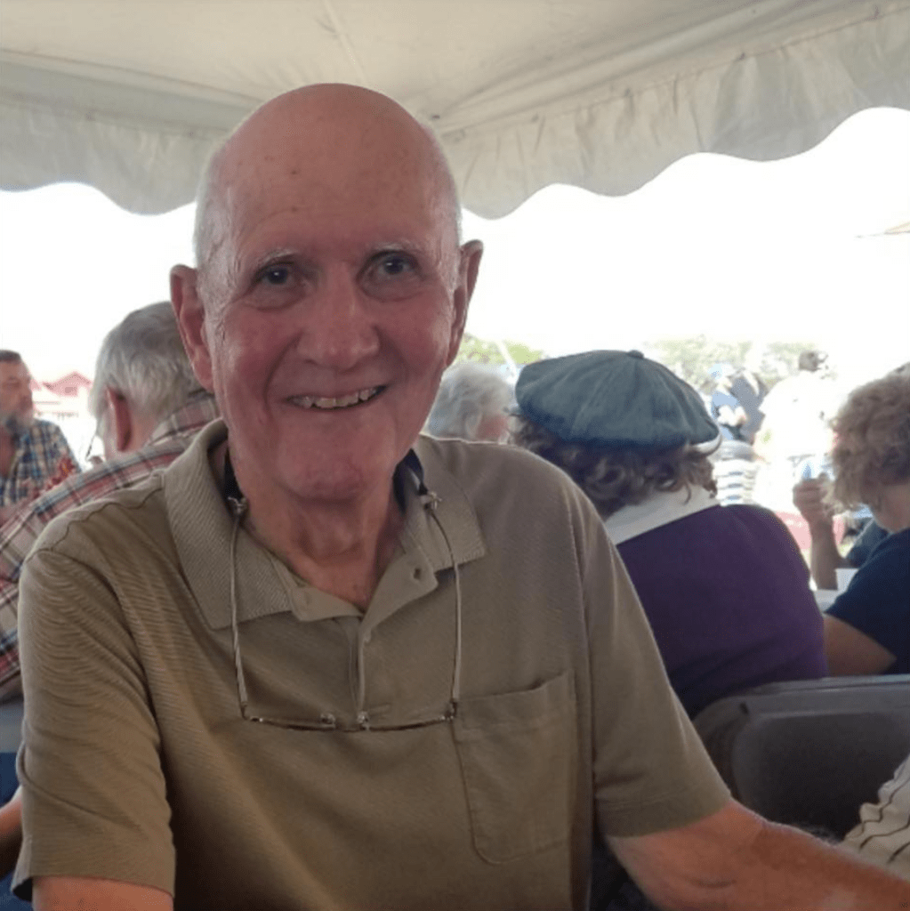 A recent photo of Bill Hassinger. He's sitting outside at a table with others and is smiling.