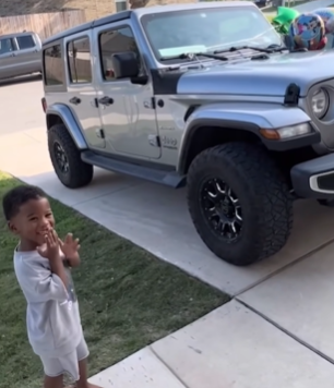 A little boy stands outside near a car. He's smiling wide and giggling as he covers his mouth with his hand. Attached to the car are birthday balloons.