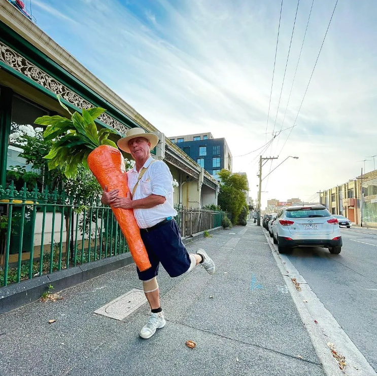 The carrot man of Melbourne poses with one leg bent and a silly smile on his face as he carries his giant carrot.