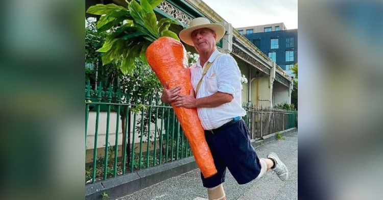The carrot man of Melbourne poses with one leg bent and a silly smile on his face as he carries his giant carrot.
