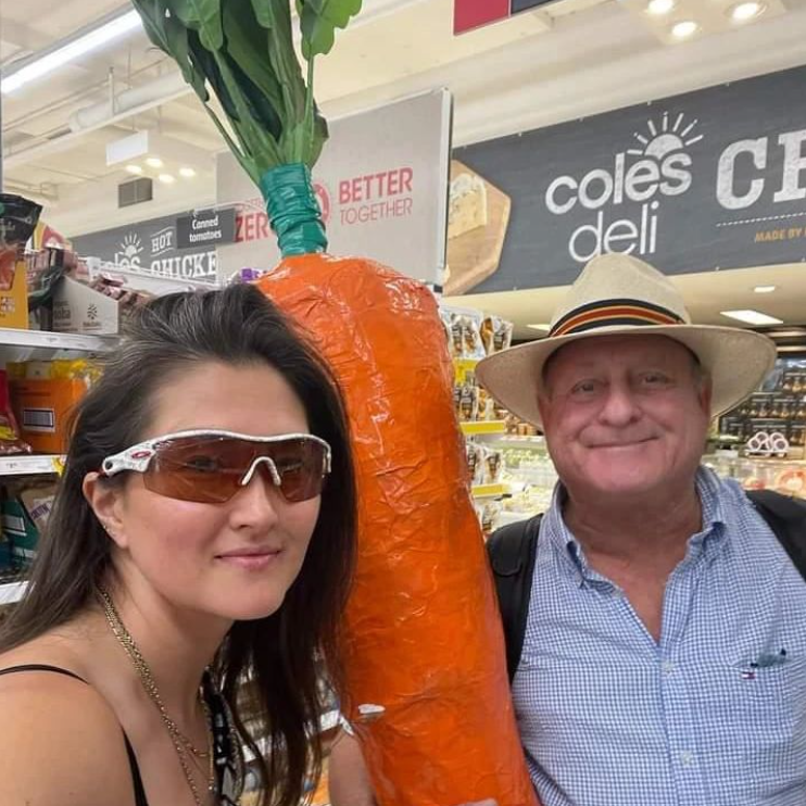 The carrot man of Melbourne smiles for a selfie with someone inside a grocery store, his giant carrot in tow.