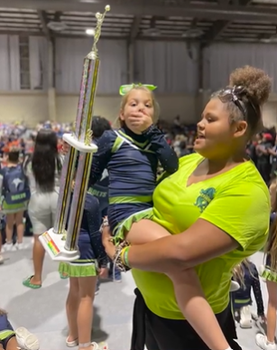 Peyton is being held in the arms of an adult. She holds her trophy in one hand and covers her mouth from shock with the other.