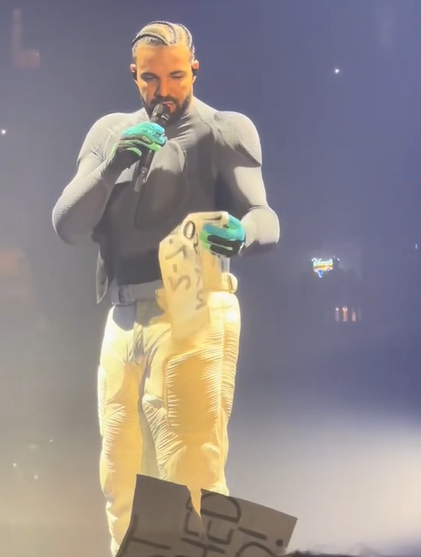 While on stage in Nashville, Drake talks into a mic as he looks down at the fan-made sign in his hand.