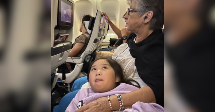 A little girl rests her head on the grandma sitting next to her on a plane. The girl is laying on her back and is looking up toward the grandma.
