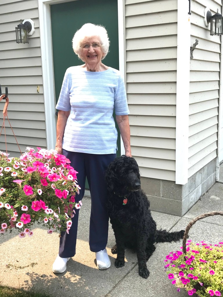 A recent photo of Joanne Blakkan. She's standing outside near a door. She's next to pink flowers and a fluffy black dog.