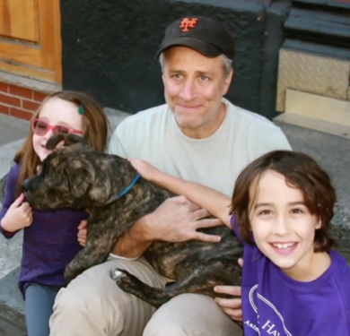 Jon Stewart and his two kids sit on a sidewalk, smiling, as they pet and hold on to their dog, Dipper.