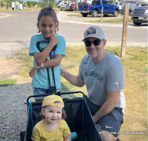 Adam Jorgenson, Charley, and Autumn pose for a photo outside. Autumn sits in a cart that can be pulled, Charley stands behind her, and Adam squats next to them. They're all smiling.