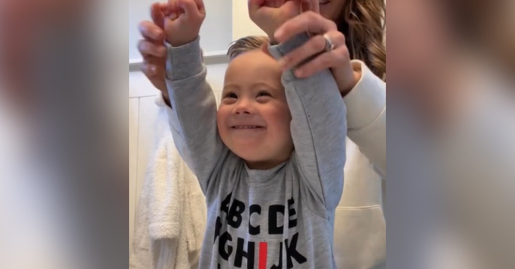 A toddler with Down syndrome smiles wide as he lifts his hands into the air. Mom stands behind him and holds onto his wrists, helping him lift his arms in celebration.