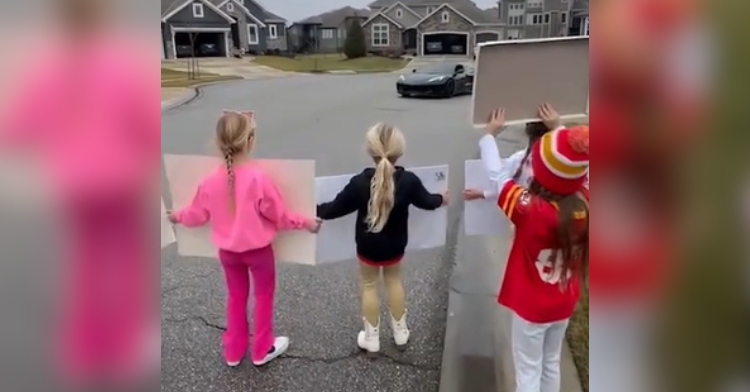 View from behind of three little kids holding up signs as they stand on the road in their neighborhood. They're giving Rashee Rice a sweet send-off to the Super Bowl.