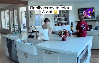 View from behind of Caitlin Teal's sons sitting on the kitchen island counter as they eat breakfast and watch the living room TV.