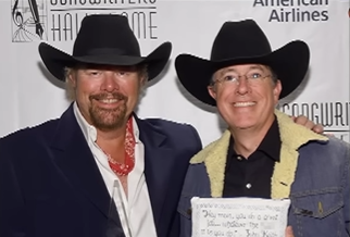 Toby Keith and Stephen Colbert smile as they pose for a photo on a red carpet. They're both wearing cowboy hats.