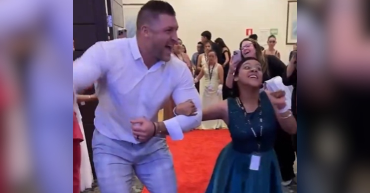 Tim Tebow smiles as he walks arm in arm with a young woman at the first Night to Shine event in 2024.