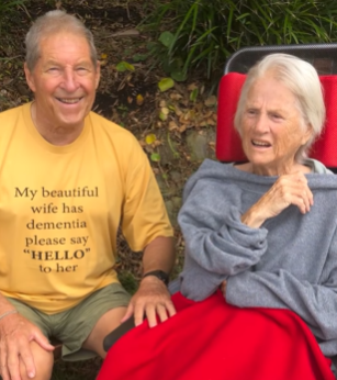Present-day Jim and Maureen: They're both sitting in their own chairs outside. Jim smiles with a hand placed on the arm rest of Maureen's chair. He's wearing a yellow t-shirt that reads: My beautiful wife has dementia please say "HELLO" to her.