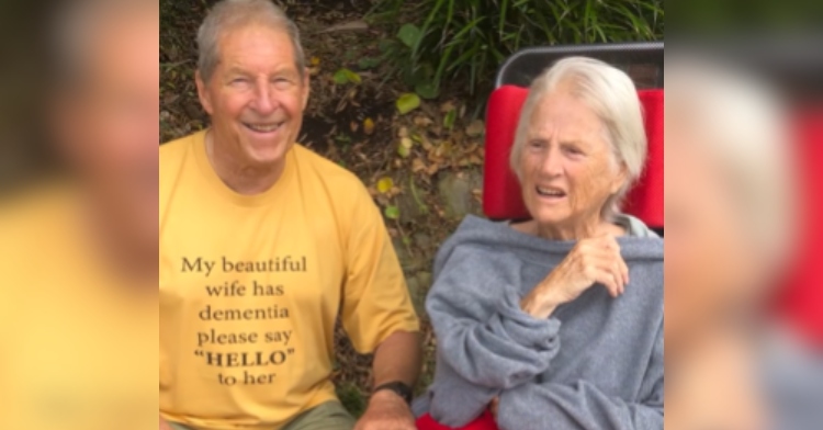 Present-day Jim and Maureen: They're both sitting in their own chairs outside. Jim smiles with a hand placed on the arm rest of Maureen's chair. He's wearing a yellow t-shirt that reads: My beautiful wife has dementia please say "HELLO" to her.