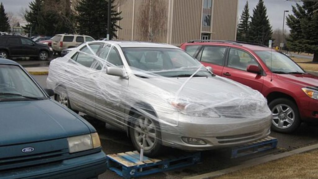 Image shows a car in a parking lot, with all four wheels on pallets and the body wrapped in shrink wrap.