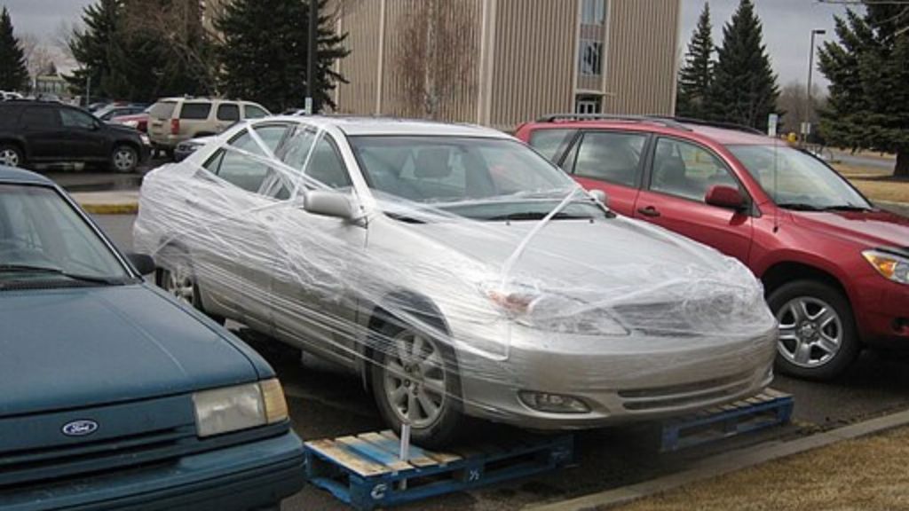 Image shows a car in a parking lot, with all four wheels on pallets and the body wrapped in shrink wrap.
