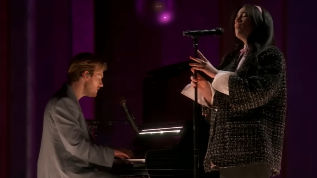 Side view of Finneas playing a piano and Billie Eilish singing into a mic as she stand near him.