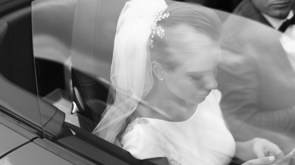 A black and white photo of a bride sitting in a car. Her groom can partially be seen in the driver's seat. She's looking at the phone in her hands.