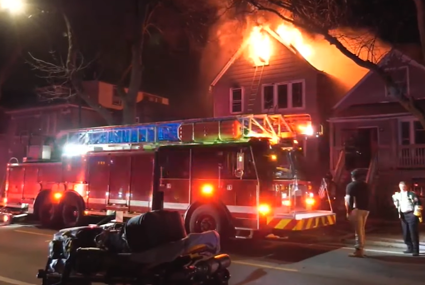 View of a home in Chicago on fire. A fire truck is just outside and there are people walking around.