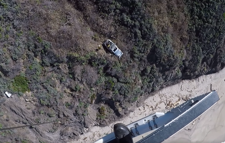 View from someone on a rescue chopper. Looking down, we see a stranded car on a cliffside.