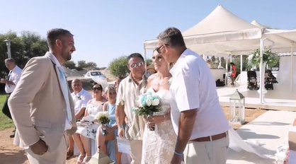 A bride, Holly Hatch, smiles as her dad and stepdad stand on either side of her. Her groom, Alfie, stands in front of them, smiling.