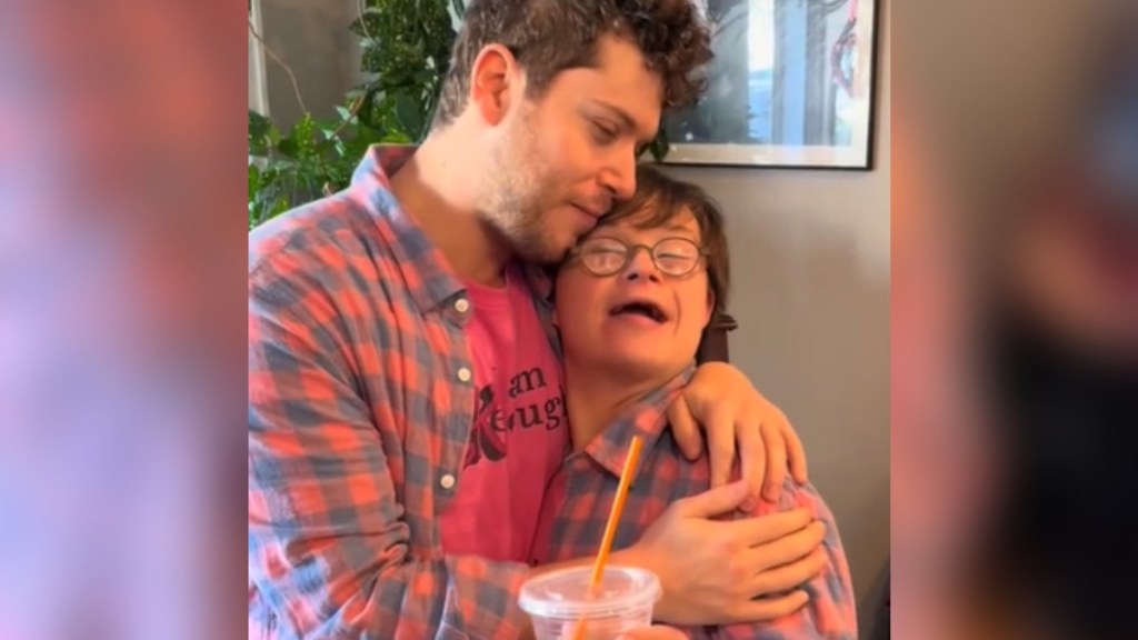 Nick and his younger brother, Gabe, hug. They're both wearing checkered pink and gray button-ups with an "I am Kenough" shirt underneath. Gabe is also holding an iced coffee.