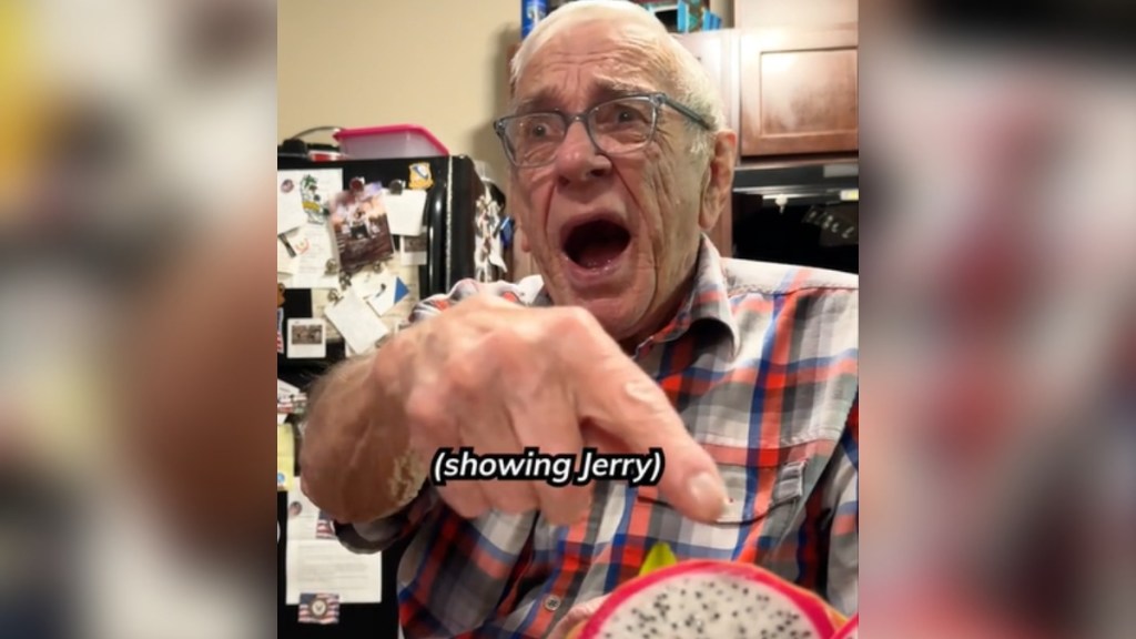 An 81-year-old man points at a dragon fruit just out of view of the camera. His eyes are wide and his mouth is open with shock and disgust. Text on the image reads: (showing Jerry)