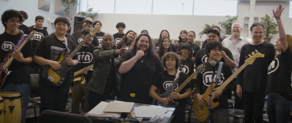Wolfgang Van Halen laughs as he poses with a large group of students with guitars as well as other adults.