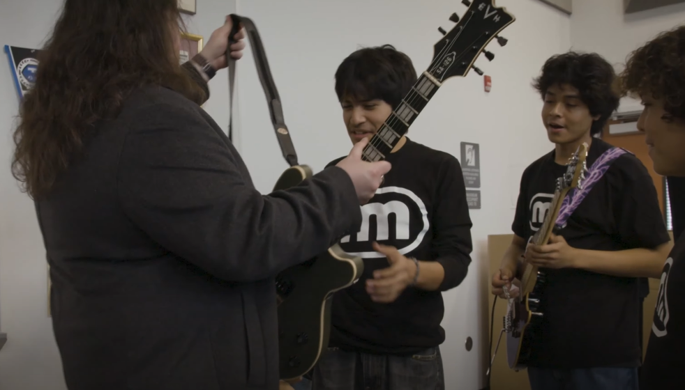 Wolfgang Van Halen hands a student a guitar. Two other students, one with his own guitar, stands by and watches.