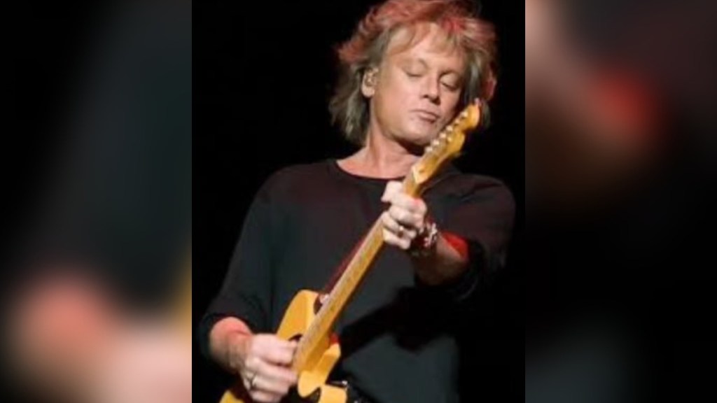 Close up of Eric Carmen looking down as he plays a guitar.