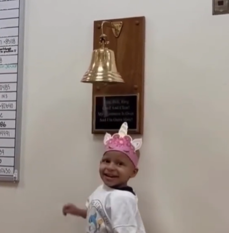 A young cancer patient wearing a pink crown and standing under a bell.