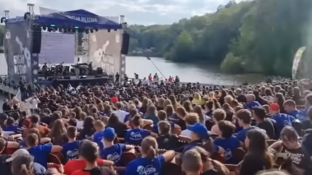 View of a crowd gathered near water as they play guitars. They're sat near a stage that's set up over the water.
