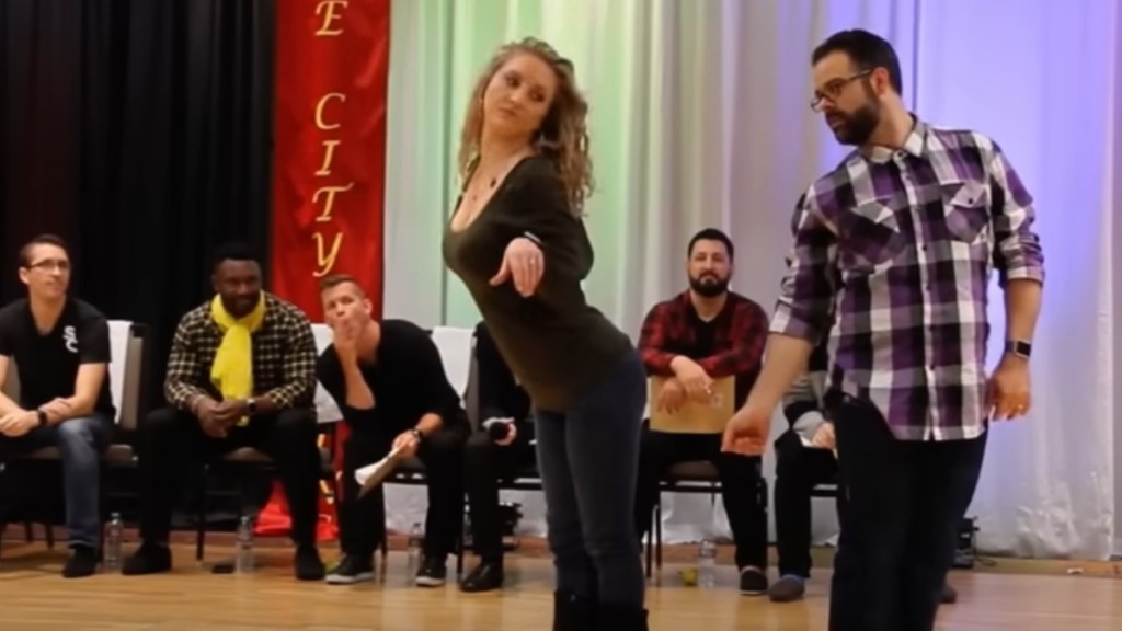 A woman and man perform an improvised dance as people sit in chairs behind them, shocked.
