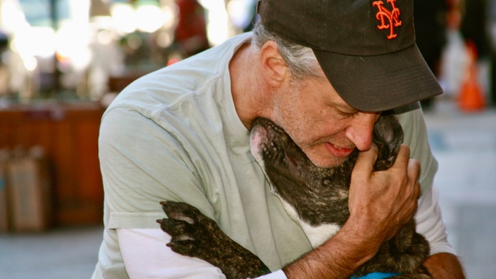 Jon Stewart holds his dog, Dipper, close for a hug. The dog buries his face into Stewart's neck and his paw rests on one of his arms like a hug.