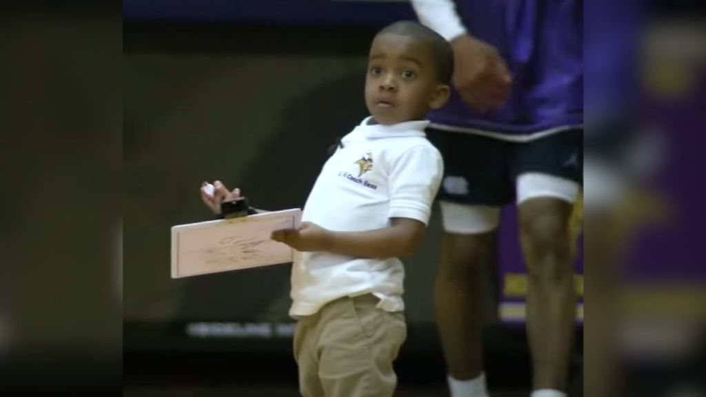 A small child holds a clipboard on the sidelines of a basketball court.
